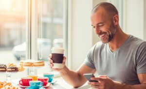 Ein Mann in einem Coffee-Shop hält einen Becher Kaffee in der Hand und blickt dabei lächelnd auf sein Smartphone. KI-generiert.